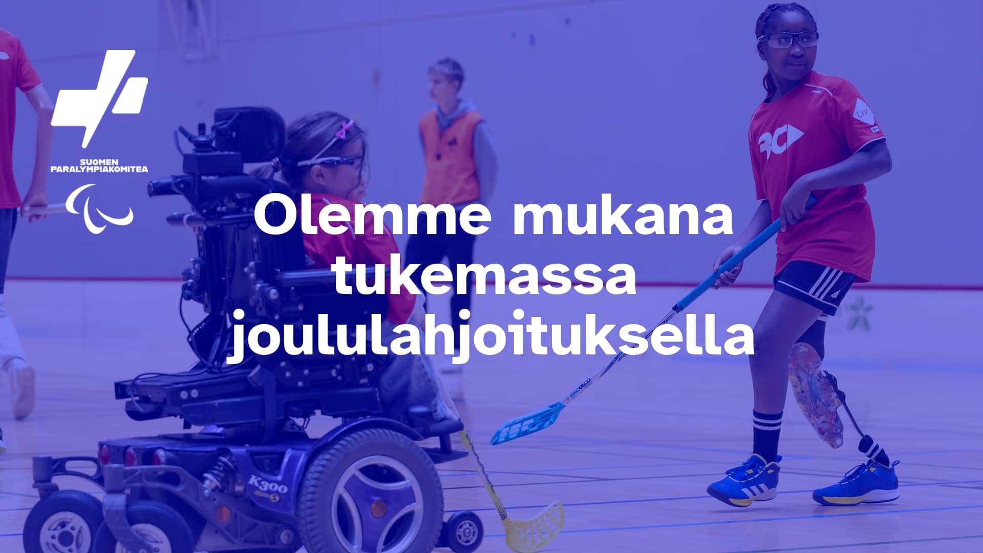 Kuva, jossa liikuntahallissa pelaa lapsia. Yksi lapsista käyttää sähkökäyttöistä pyörätuolia ja toinen liikuntaproteesia. Heillä on salibandymailat käsissään. Kuvassa näkyy myös Suomen Paralympiakomitean logo sekä teksti “Olemme mukana tukemassa joululahjoituksella”.
