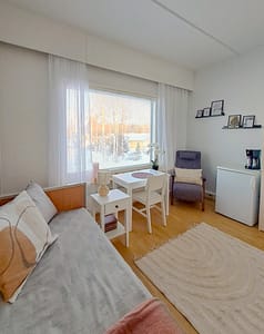 Asukashuone, jossa sänky, pöytä ikkunan äärellä, nojatuoli ja pieni jääkaappi.