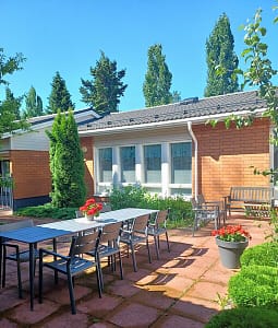 Hoivakodin rakennus ulkoa, vehreässä ympäristössä ja edustalla terassikalusteita ja kukkia.