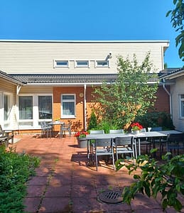 Hoivakodin rakennus ulkoa, vehreässä ympäristössä ja edustalla terassikalusteita ja kukkia.