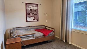 Kuva hoivakodin huoneesta, jossa ikkunan vieressä on sänky, yöpöytä ja ryijy seinällä.