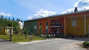 Hoivakodin vihreä ja oranssi rakennusosa, parveke kiertoportaineen, aidan vieressä tuolirivi ja polkupyöriä.