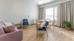 Valoisa oleskelu- ja ruokailutila, jossa tuoleja, pöytä ja suuret ikkunat hoivakodissa.
