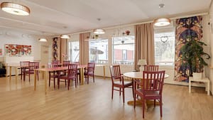 Valoisa ruokailutila, pöytiä ja tuoleja sekä suuret ikkunat.