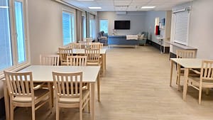 Valoisa ruokailu- ja oleskelutila hoivakodissa, pöytiä, tuoleja ja tv-alue suurten ikkunoiden äärellä.