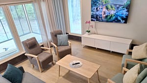 Rauhallinen oleskelutila hoivakodissa, nojatuoleja, sohva ja tv vaaleassa ympäristössä.