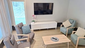 Kodikas oleskelunurkkaus hoivakodissa, nojatuoleja, sohva ja televisio luonnonvaloisassa tilassa.