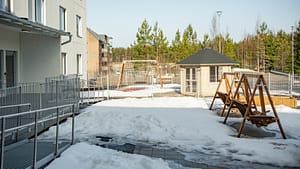 Hoivakodin piha-alue talvella, jossa esteetön kulku, keinut ja pieni piharakennus.
