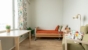 Asukashuone, jossa säädettävä hoivasänky, pöytä, kasveja ja värikkäät tekstiilit tuomassa kodikkuutta.