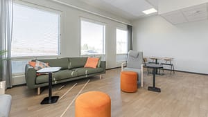 Valoisa oleskelutila, jossa sohva, nojatuoli ja pieniä pöytiä suurten ikkunoiden äärellä.