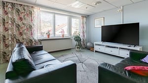 Valoisa oleskelutila, jossa sohva, sohvapöytä, kirjahylly ja suuret ikkunat verhoineen.