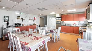 Tilava keittiö ja ruokailutila, useita pöytiä ja tuoleja, kodinkoneet ja kaapistot.