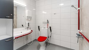 Esteetön kylpyhuone, jossa punainen suihkutuoli, tukikahvat ja wc.