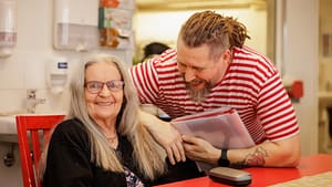 Asukas ja hoitaja istuvat sängyllä punertavan ryijyn edessä ja katsovat hymyillen kameraan.