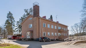 Kaksikerroksinen vaalea asuinrakennus, piha-alue ja pysäköityjä autoja.
