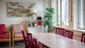 Hoivakodin valoisa huone, jossa on pitkä pöytäryhmä, nojatuoleja, kasveja ja muita sisustuselementtejä ikkunan edessä.