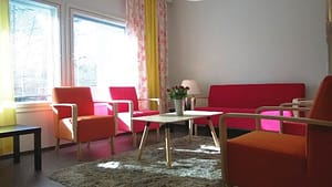Hoivakodin olohuone, jossa on punaisia huonekaluja (sohva, tuoleja) valoisan ikkunan edustalla.