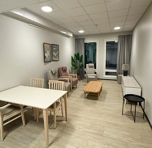 Kodikas oleskelutila, jossa kolme nojatuolia, puinen sohvapöytä, ruokapöytä ja suuret ikkunat.