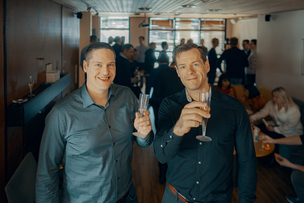 Two men standing side by side at a party and raising champagne glasses.