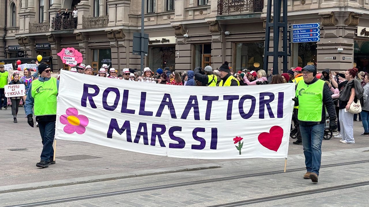 Kaksi henkilöä kulkee marssivan joukon ensimmäisinä välissään rollaattorimarssin banderolli