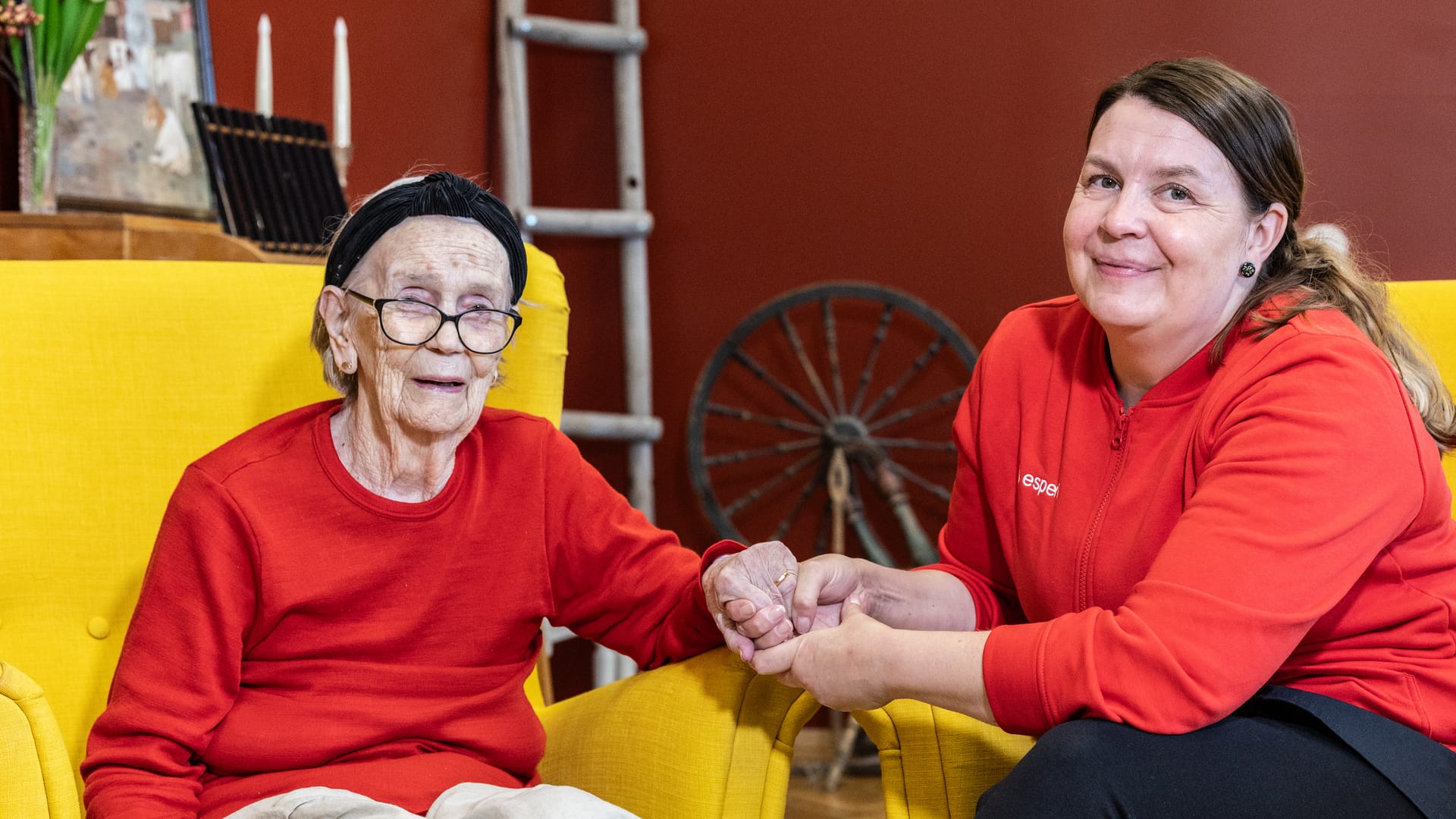 Ikääntynyt asukas ja hoitaja istuvat nojatuoleilla ja pitävät käsistä kiinni.