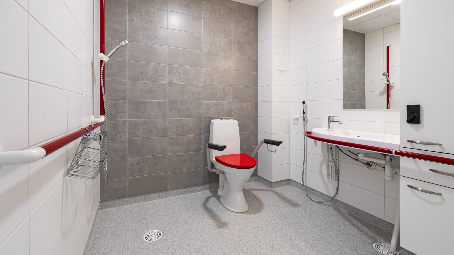 Esteetön kylpyhuone hoivakodissa, jossa tukikaiteet, suihku ja korotettu wc-istuin punaisin yksityiskohdin.